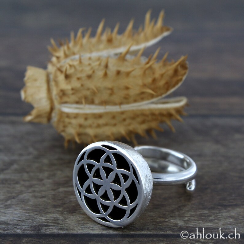 May include: A silver ring with a round, openwork design featuring a flower-like pattern. The ring is set against a wooden background.