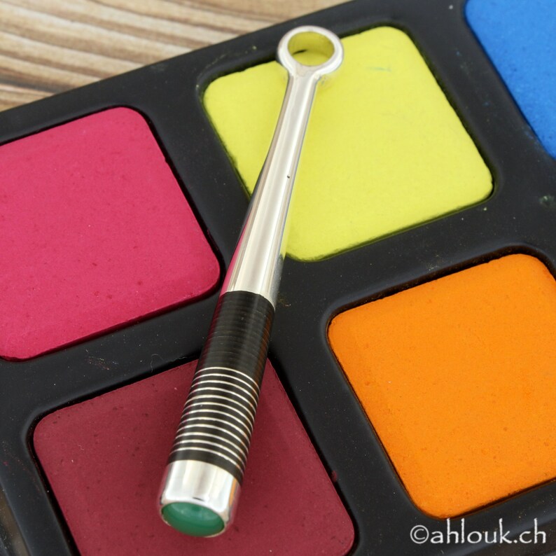 May include: A silver and black metal stick with a green stone at the end. The stick is lying on a black and white surface with colorful squares.