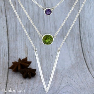 May include: A silver necklace with three pendants. The top pendant is a small purple gemstone, the middle pendant is a larger green gemstone, and the bottom pendant is a long, thin, silver V-shape.