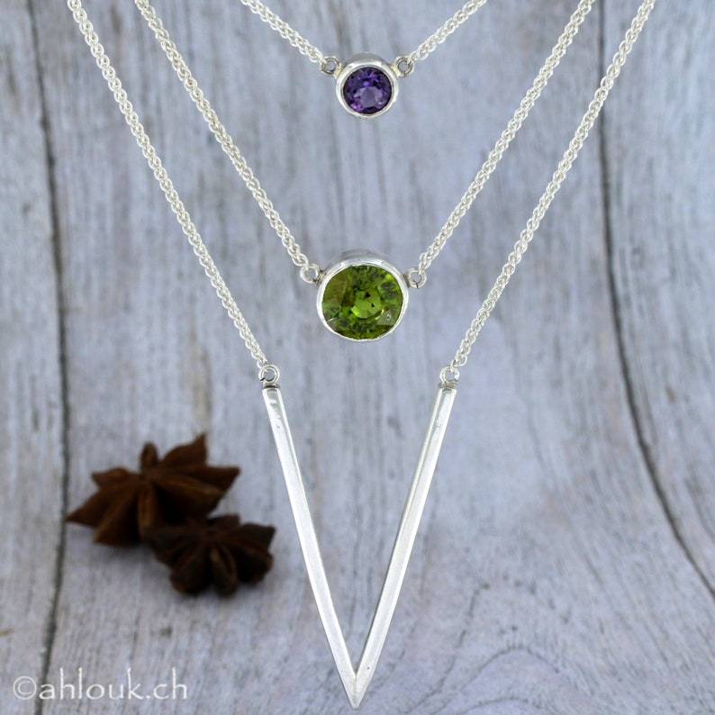 May include: A silver necklace with three pendants. The top pendant is a small purple gemstone, the middle pendant is a larger green gemstone, and the bottom pendant is a long, thin, silver V-shape.