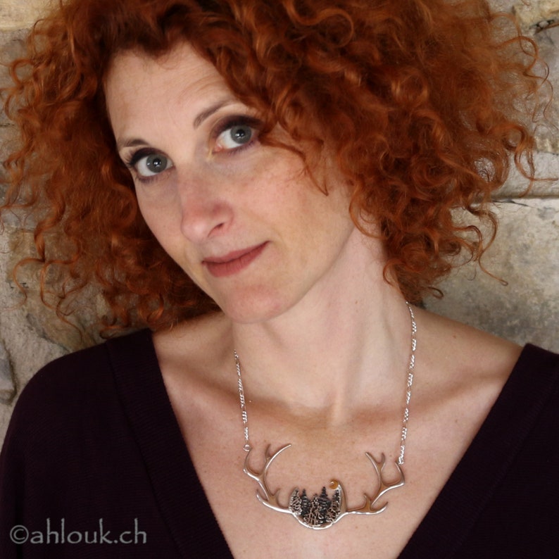May include: A woman with long, curly red hair is wearing a silver necklace with a pendant shaped like a deer's antlers. The pendant features a detailed design of a forest scene with trees and a moon.
