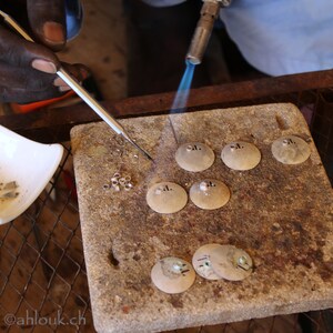 Puede incluir: Una persona utiliza un soplete para calentar peque&ntilde;as piezas de metal redondas sobre una superficie de piedra. Las piezas de metal tienen peque&ntilde;os agujeros en el centro. Tambi&eacute;n hay peque&ntilde;os anillos de metal en la superficie.