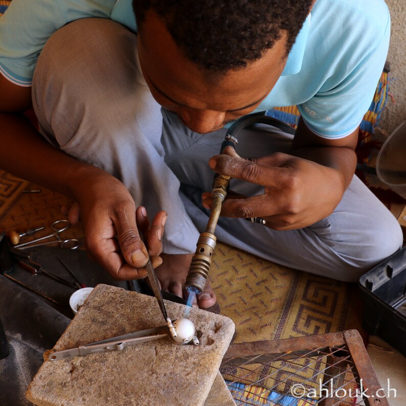 May include: A person uses a torch to heat a small, white pearl while holding a pair of tweezers. The pearl is on a gray stone surface.