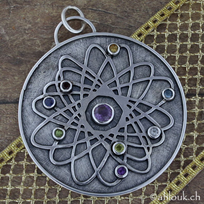 May include: A silver pendant with a circular design featuring a central amethyst gemstone surrounded by smaller gemstones in various colors, including green, blue, and yellow. The pendant has a delicate, intricate pattern that resembles a stylized atom.
