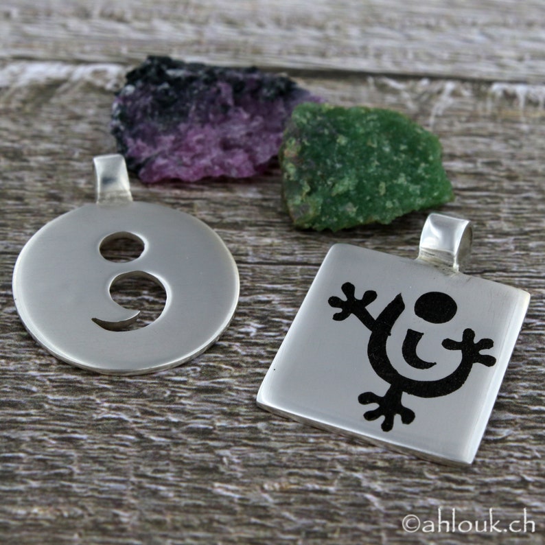May include: Two silver pendants with black designs. One pendant is round with a semicolon symbol cut out. The other pendant is square with a stylized figure with a circle in the center.