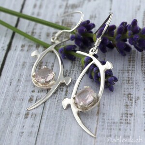 Pu&ograve; includere: Un paio di orecchini d'argento con un design delicato e astratto. Ogni orecchino presenta una pietra preziosa rosa incastonata in una cornice d'argento.