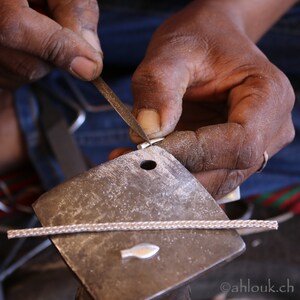 May include: A person uses a file to shape a silver pendant on a metal anvil. The pendant is shaped like a fish.