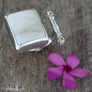 May include: A silver ring with a white, rectangular stone set in the band. The ring is open and has a silver bar with a decorative design on the end.