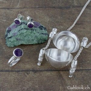 May include: A set of silver jewelry featuring a ring, earrings, and a pendant necklace. The ring and earrings have purple gemstones. The pendant is a silver sphere with a hinged lid.