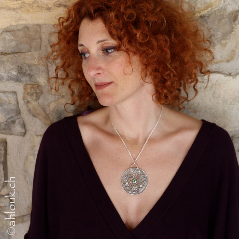 May include: A woman with red curly hair wearing a dark purple top and a silver necklace with a large circular pendant featuring a green gemstone surrounded by smaller stones.