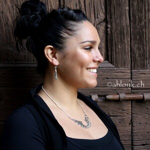 May include: A woman wearing a black top with a silver necklace and earrings. The necklace features a geometric design with small green stones. The earrings are long and silver with a geometric design.