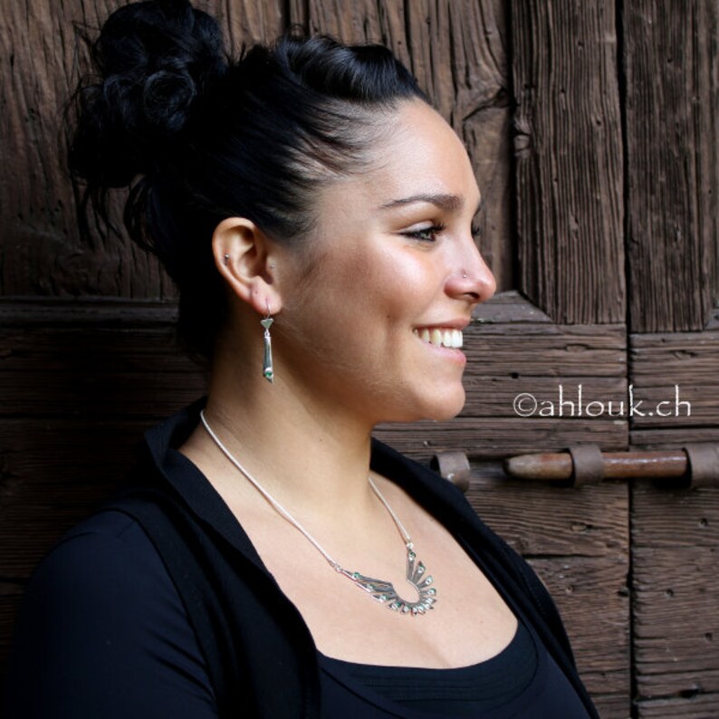 May include: A woman wearing a black top with a silver necklace and earrings. The necklace features a geometric design with small green stones. The earrings are long and silver with a geometric design.
