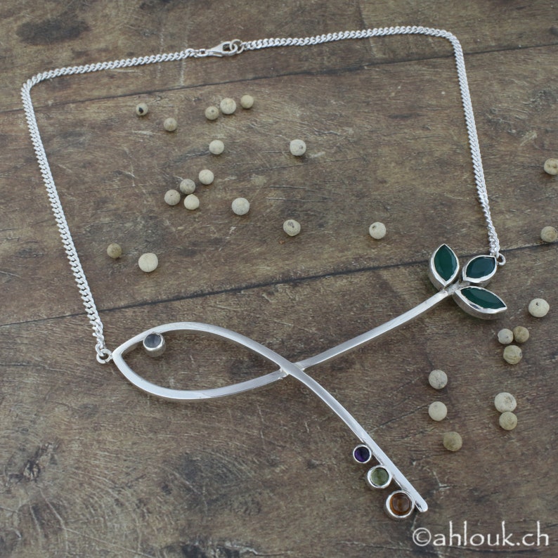 May include: A silver necklace with a delicate chain and a pendant shaped like a stylized fish with a small blue gemstone. The pendant is connected to a long, thin stem with a cluster of green gemstones at the top, resembling leaves.