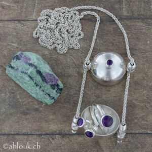 May include: A silver necklace with a pendant that holds three rings with purple gemstones. The necklace is on a wooden surface with a green and black stone.