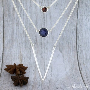 May include: A silver necklace with three chains and three gemstone pendants. The top pendant is a small red gemstone, the middle pendant is a larger blue gemstone, and the bottom pendant is a silver V-shaped pendant.