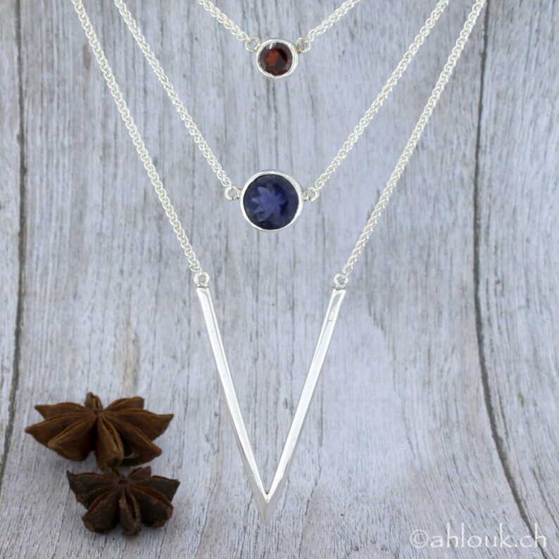 May include: A silver necklace with three chains and three gemstone pendants. The top pendant is a small red gemstone, the middle pendant is a larger blue gemstone, and the bottom pendant is a silver V-shaped pendant.