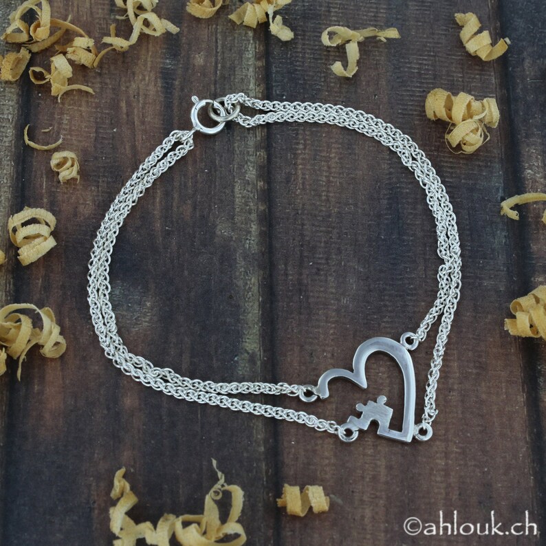 May include: A silver chain bracelet with a heart-shaped charm featuring a puzzle piece design. The bracelet is double-stranded and has a lobster clasp closure.