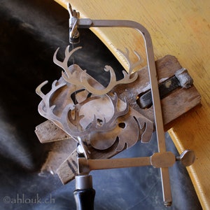 May include: A silver metal saw with a black handle is used to cut out a metal reindeer design. The reindeer is being cut out on a wooden board.