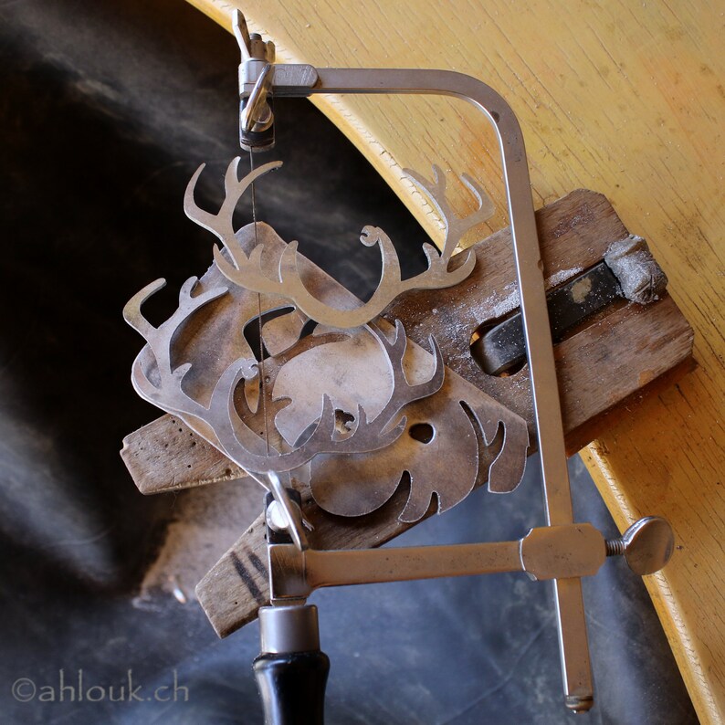 May include: A silver metal saw with a black handle is used to cut out a metal reindeer design. The reindeer is being cut out on a wooden board.