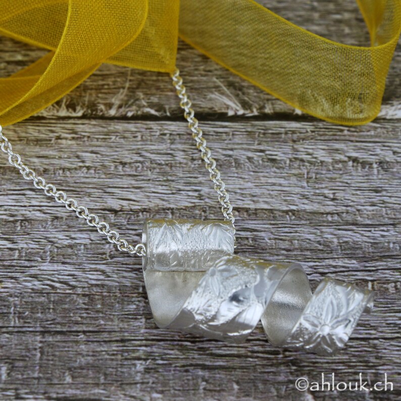 May include: A silver necklace with a textured, twisted pendant. The pendant is shaped like a ribbon or scroll. The necklace is on a wooden surface with a yellow ribbon.