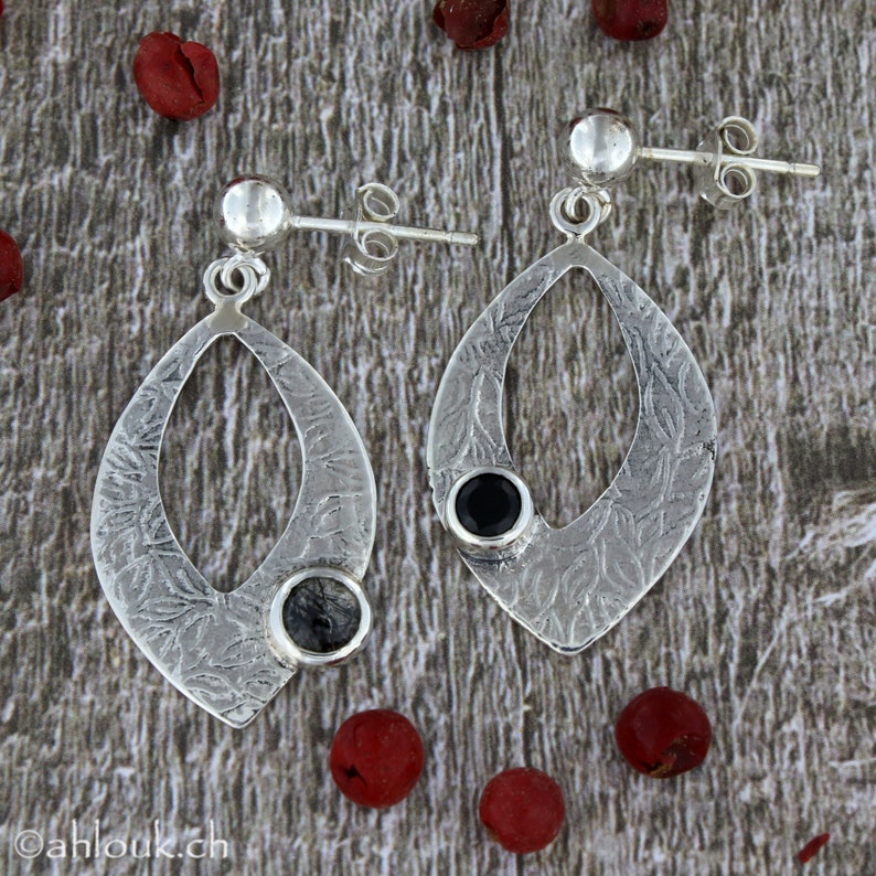 May include: A pair of silver earrings with a textured oval design. Each earring features a black gemstone and a smaller clear gemstone.