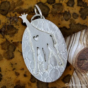 May include: A silver pendant with an oval shape and a giraffe design. The giraffe has a heart-shaped cutout on its body. The pendant is suspended from a silver loop. The background is a textured, brown surface.