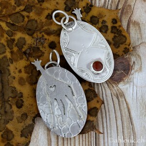 May include: Two silver oval pendants with giraffe designs. One pendant features a giraffe silhouette with a heart cutout, the other has geometric patterns and a red stone. Both have a giraffe head at the top and are on a leaf.
