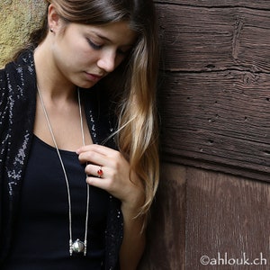 May include: A woman wearing a black top with a silver necklace and a red gemstone ring. The necklace features a silver pendant with a round, textured design.