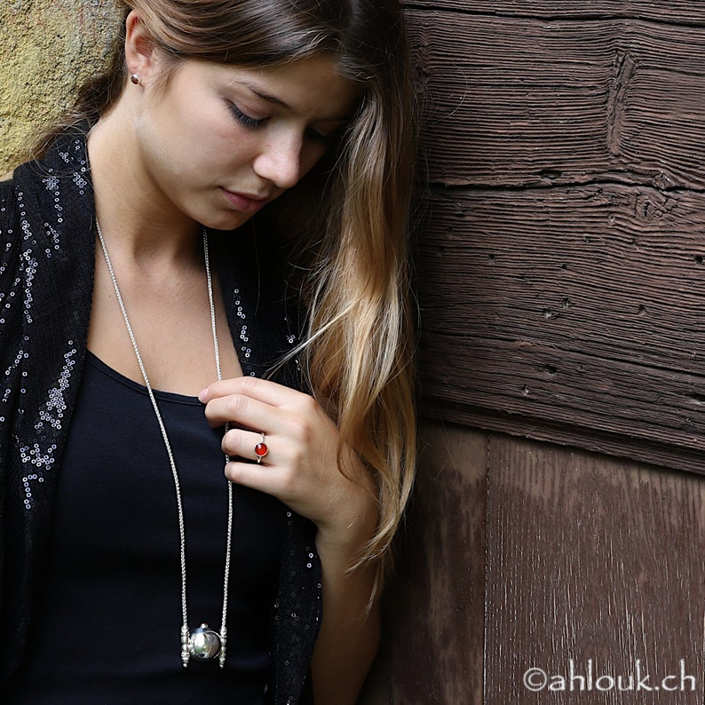 May include: A woman wearing a black top with a silver necklace and a red gemstone ring. The necklace features a silver pendant with a round, textured design.
