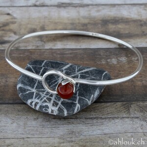 May include: A silver bangle bracelet with a red carnelian stone accent. The bangle is open and has a simple design.