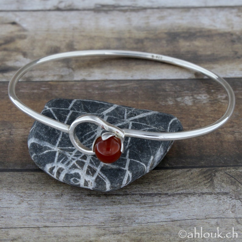 May include: A silver bangle bracelet with a red carnelian stone accent. The bangle is open and has a simple design.