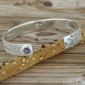 Puede incluir: Pulsera de plata con un dise&ntilde;o tejido y dos piedras redondas. Una piedra es azul y la otra es azul claro.