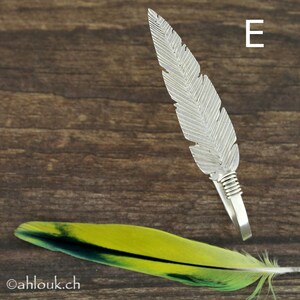 May include: A silver feather ring with a textured design. The ring is on a wooden surface with a green and yellow feather in the foreground.