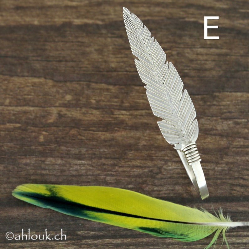 May include: A silver feather ring with a textured design. The ring is on a wooden surface with a green and yellow feather in the foreground.