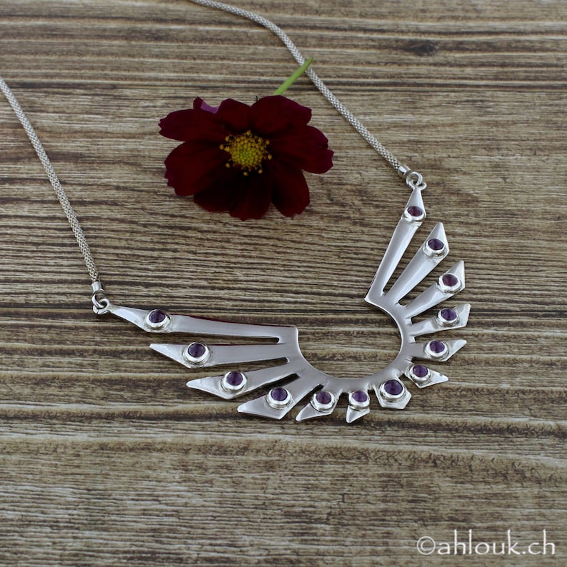 May include: A silver necklace with a geometric design featuring amethyst gemstones. The necklace is in the shape of wings with a rounded bottom.
