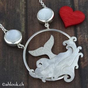 May include: Silver necklace with a whale tail pendant and two white moonstone beads. The whale tail is surrounded by a circle of silver waves.