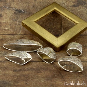 May include: A collection of silver-toned jewelry pieces with textured surfaces, including oval and ring-shaped designs. A gold-colored, diamond-shaped picture frame is in the background. The jewelry is displayed on a wooden surface.