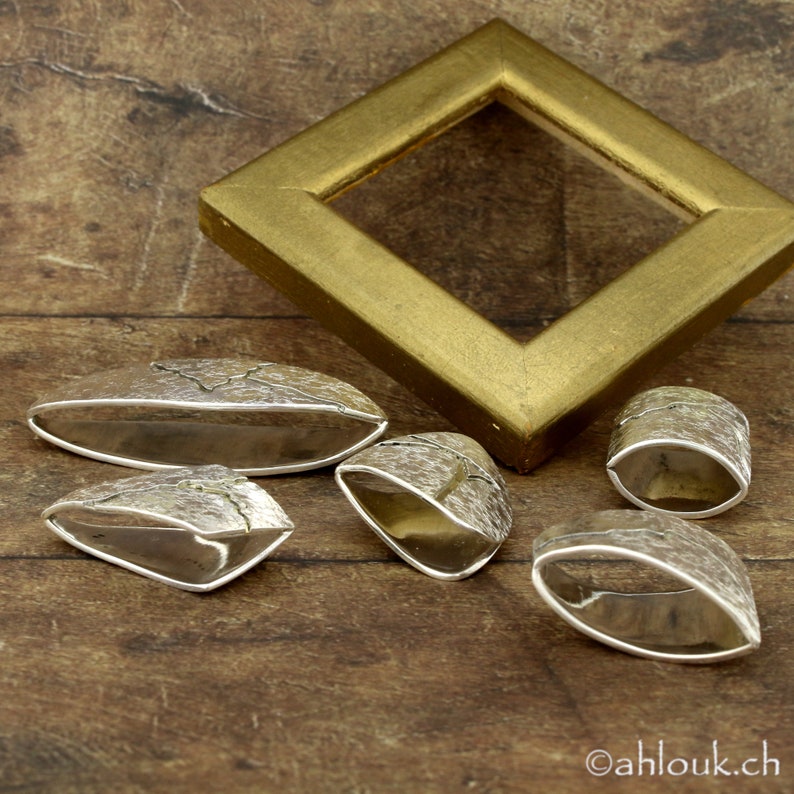 May include: A collection of silver-toned jewelry pieces with textured surfaces, including oval and ring-shaped designs. A gold-colored, diamond-shaped picture frame is in the background. The jewelry is displayed on a wooden surface.