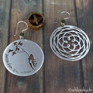 May include: A pair of silver earrings with a delicate design. One earring features a cutout of a girl flying with birds and the text "I know the way home". The other earring has a circular design with a repeating pattern.
