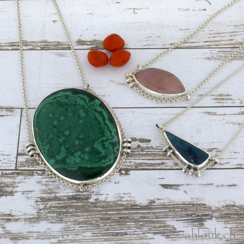 Puede incluir: Tres collares de plata con diferentes colgantes de piedras preciosas. El primer collar tiene un gran colgante ovalado de malaquita verde. El segundo collar tiene un colgante ovalado rosa de cuarzo rosa. El tercer collar tiene un colgante triangular azul.