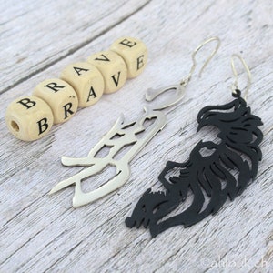 May include: A pair of earrings with a black and silver design. The earrings are shaped like a stylized bird with a wingspan. The word "BRAVE" is spelled out in wooden blocks.