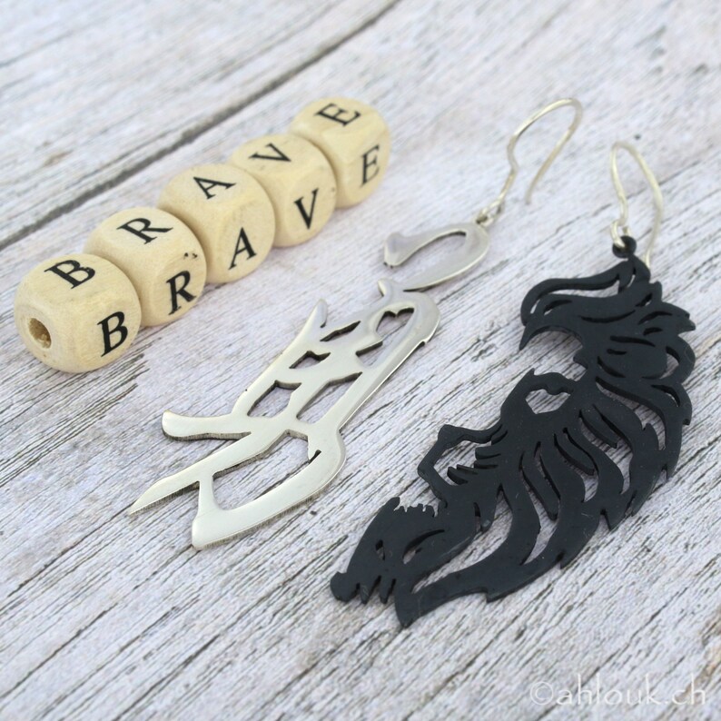 May include: A pair of earrings with a black and silver design. The earrings are shaped like a stylized bird with a wingspan. The word "BRAVE" is spelled out in wooden blocks.