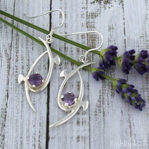 Pu&ograve; includere: Un paio di orecchini d'argento con un design delicato e astratto. Ogni orecchino presenta una pietra preziosa di ametista viola incastonata in una cornice d'argento. Gli orecchini sono appesi a un gancio d'argento.
