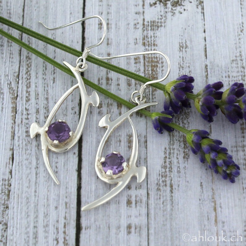 Pu&ograve; includere: Un paio di orecchini d'argento con un design delicato e astratto. Ogni orecchino presenta una pietra preziosa di ametista viola incastonata in una cornice d'argento. Gli orecchini sono appesi a un gancio d'argento.