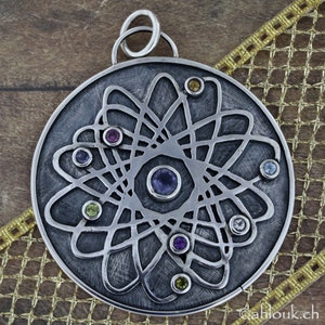 May include: A silver pendant with a circular design featuring a stylized atom with a central nucleus and orbiting electrons. The pendant is adorned with colorful gemstones in shades of purple, green, yellow, and blue.