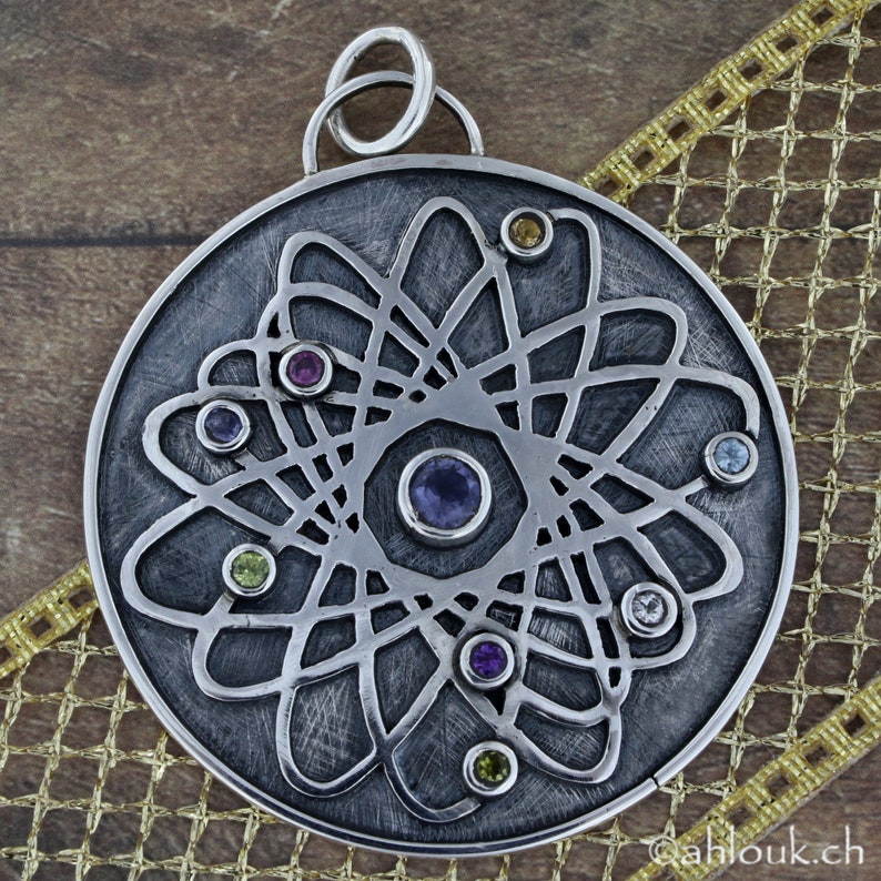 May include: A silver pendant with a circular design featuring a stylized atom with a central nucleus and orbiting electrons. The pendant is adorned with colorful gemstones in shades of purple, green, yellow, and blue.