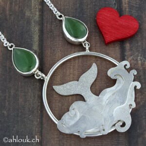 May include: A silver necklace with a whale tail pendant and two green jade drops. The pendant is a circle with a whale tail and waves cut out of the metal. The necklace is on a wooden surface.