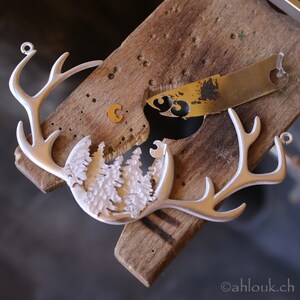 May include: A silver metal deer head ornament with a detailed design of a forest scene in the center. The ornament is being worked on with tools on a wooden surface.