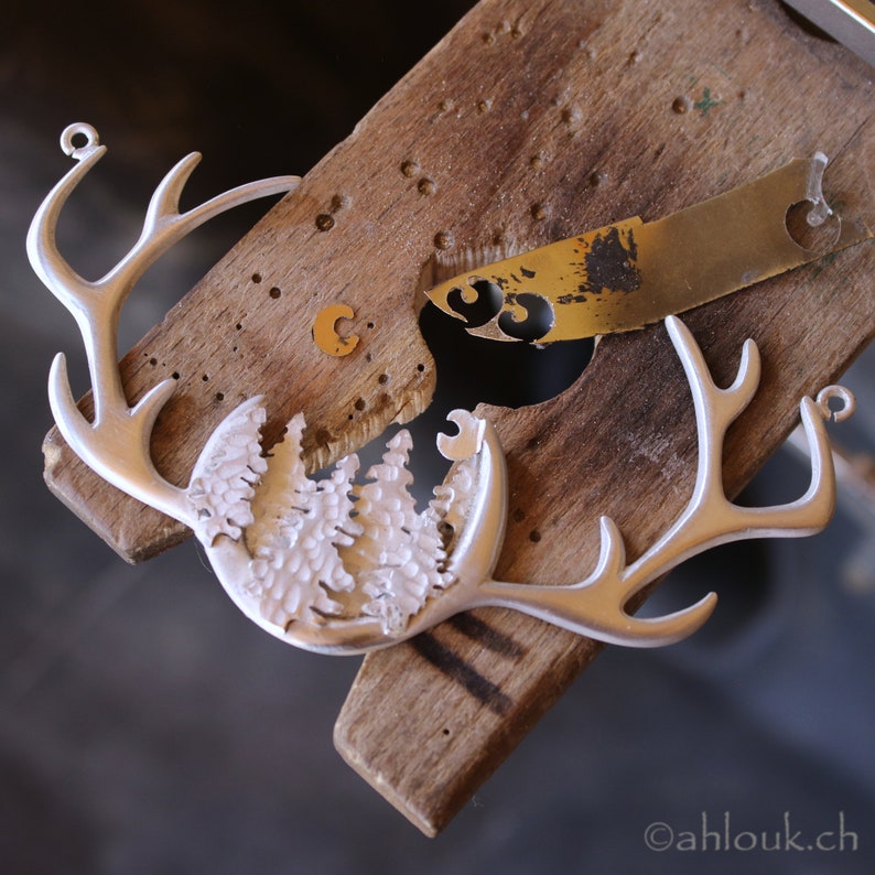 May include: A silver metal deer head ornament with a detailed design of a forest scene in the center. The ornament is being worked on with tools on a wooden surface.