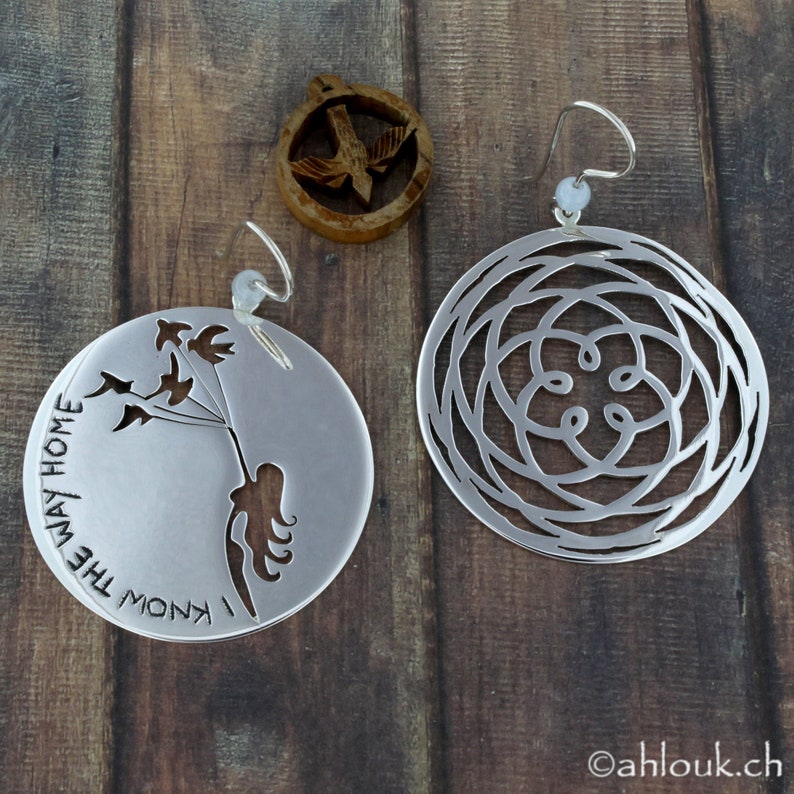 May include: A pair of silver earrings. One earring features a circular design with a cutout pattern. The other earring features a cutout design of a person holding birds with the text "I know the way home".